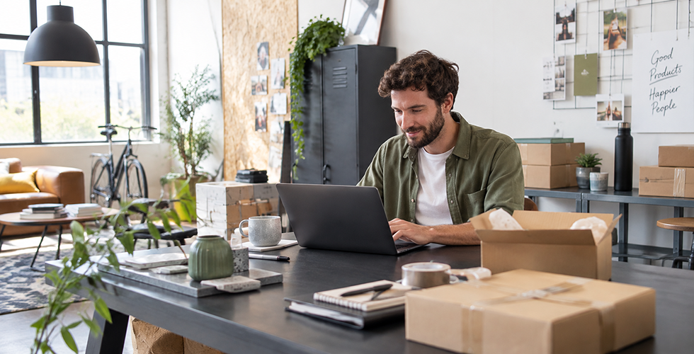Junger Mann arbeitet in einem kreativen Loftbüro an einem Laptop, umgeben von Versandkartons und Verpackungsmaterial.